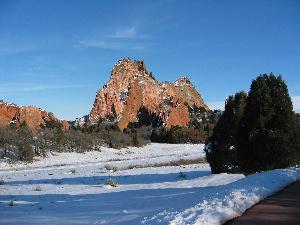 A snow-covered landscape featuring rocky formations and a clear blue sky. In the foreground, there's a pathway winding through the snow, with pine trees and a field visible in the background. The majestic rock formations rise prominently against the sky. Garden of the Gods: Ute Trail mountain bike trail.