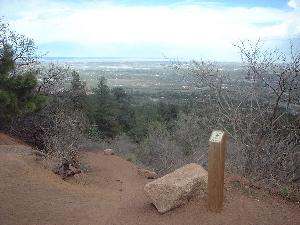 A scenic view from a mountain trail, featuring a rocky pathway, sparse vegetation, and a wooden post marked with information. In the background, a panoramic vista of rolling hills and valleys under a cloudy sky is visible. Stratton Open Space / The Chutes mountain bike trail.