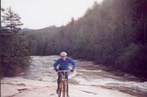 A person in a blue sweater and helmet is riding a mountain bike along a rocky riverbank, with a forest of trees and hills in the background. The river flows rapidly beside them. DuPont State Forest mountain bike trail.