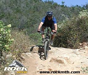 A mountain biker navigating a rocky trail surrounded by greenery on a sunny day. Sycamore Canyon mountain bike trail.