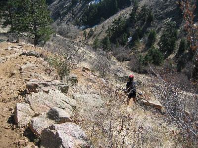 A person walking along a rocky trail in a mountainous area, surrounded by dry vegetation and trees, with a steep slope leading down to a river in the background. Apex Park mountain bike trail.