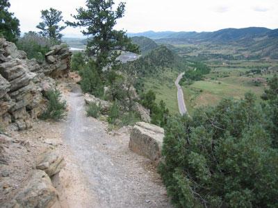 A scenic view from a rocky trail overlooking a lush valley and winding road. The landscape is dotted with trees and mountains in the distance under a cloudy sky. Red Rocks / Dakota Ridge mountain bike trail.