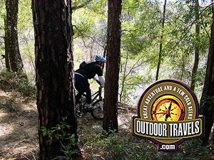 A person riding a mountain bike on a narrow trail surrounded by tall trees and greenery, with a logo featuring the text "Outdoor Travels" in the foreground. Withlacoochee State Forest: Croom Section mountain bike trail.