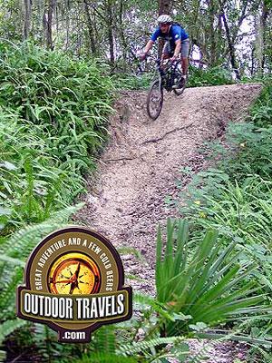 A mountain biker navigates a sandy, elevated trail surrounded by lush greenery in a forested area. The image features the logo for "Outdoor Travels" at the bottom, promoting adventure and outdoor exploration. Loyce E. Harpe Park mountain bike trail.