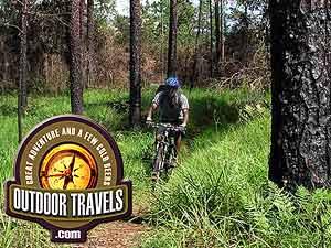 A person riding a mountain bike along a narrow trail surrounded by lush greenery and trees. An "Outdoor Travels" logo is featured in the foreground, highlighting adventure and outdoor exploration. Withlacoochee State Forest: Croom Section mountain bike trail.