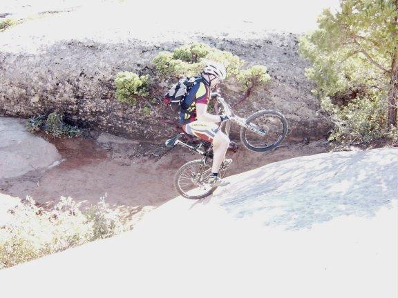 A cyclist wearing a helmet and colorful athletic gear performs a wheelie on a rocky trail, with a backdrop of greenery and rugged terrain. Gooseberry Mesa mountain bike trail.