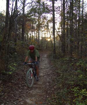 A person riding a mountain bike on a narrow dirt trail through a wooded area, with trees and fallen leaves visible. The sun is setting in the background, casting a warm glow through the foliage. Yellow River mountain bike trail.