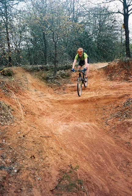 A cyclist in a green shirt and helmet riding down a dirt trail surrounded by trees, navigating a berm in a mountain biking area. The trail has a reddish-brown color, indicating off-road terrain. Lake Stanley Draper mountain bike trail.