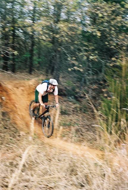 A cyclist in motion navigating a dirt trail surrounded by trees, demonstrating action and speed with a blurred background. The cyclist is wearing a helmet and sporting athletic gear, capturing the thrill of mountain biking. Lake Stanley Draper mountain bike trail.