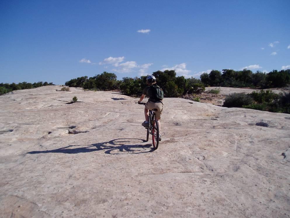 A person riding a mountain bike on a rocky, open landscape under a clear blue sky, with green shrubs in the background. Klondike Bluffs mountain bike trail.