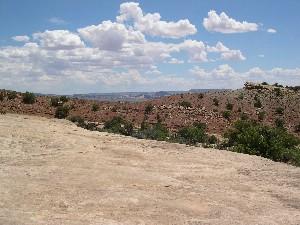 A wide view of a desert landscape featuring rocky terrain, sparse vegetation, and a blue sky with fluffy white clouds. The horizon is visible in the distance, showcasing natural formations. Klondike Bluffs mountain bike trail.