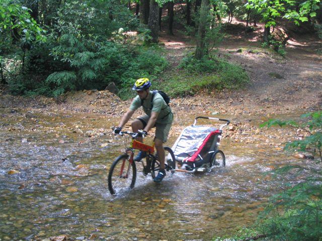 A person riding a mountain bike through a shallow stream in a forested area, pulling a bicycle trailer behind them. The scene features lush greenery and rocky terrain, with sunlight filtering through the trees. The cyclist is wearing a helmet and casual outdoor clothing. Bull / Jake Mountain mountain bike trail.