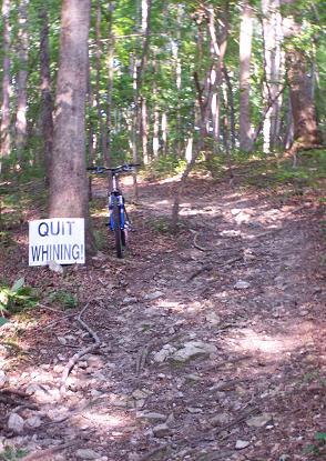 A mountain bike is parked next to a trail sign in a forested area. The sign reads "QUIT WHINING!" in bold letters, positioned on a rocky and uneven path surrounded by trees. Pee Wee's Mountain Bike Park mountain bike trail.