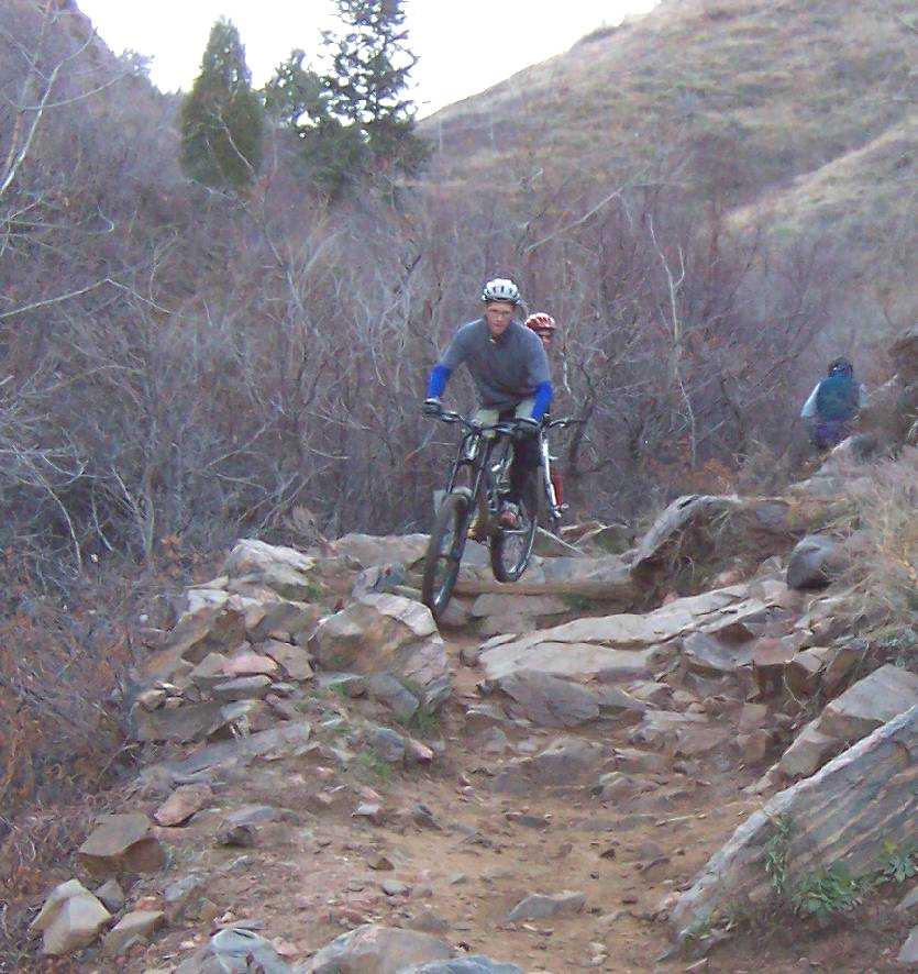 A person riding a mountain bike over rocky terrain, surrounded by sparse trees and shrubs on a natural trail. Another cyclist is visible in the background, navigating the same trail. The setting appears to be a rugged outdoor area, indicating a challenging biking course. Apex Park mountain bike trail.