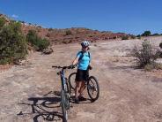 A person wearing a blue shirt and helmet stands next to a mountain bike on a rocky outdoor trail. The landscape features red rock formations and sparse vegetation under a clear blue sky. Klondike Bluffs mountain bike trail.