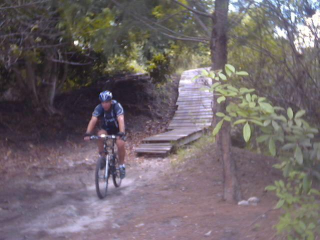 A mountain biker riding on a dirt trail near a wooden ramp surrounded by trees and foliage. Markham Park mountain bike trail.