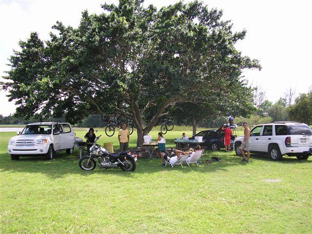 A group of people gathered under a large tree in a grassy area, enjoying a casual outing. Several vehicles, including cars and a motorcycle, are parked nearby. People are seated around a table, some are standing, and a couple of bicycles are hanging from the tree. The scene conveys a relaxed outdoor atmosphere. Markham Park mountain bike trail.