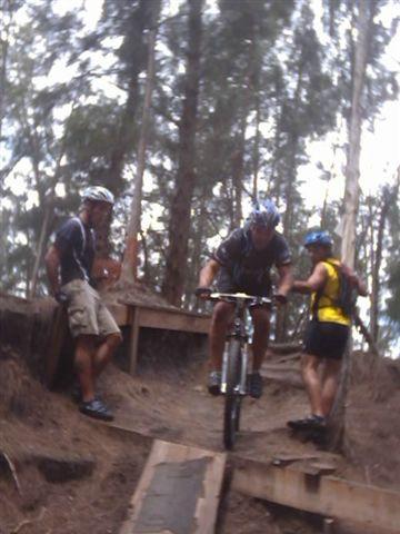 A mountain biker navigates a wooden ramp on a forest trail while two spectators watch from the side. The scene is set among tall trees, with the biker wearing a helmet and gear. Markham Park mountain bike trail.