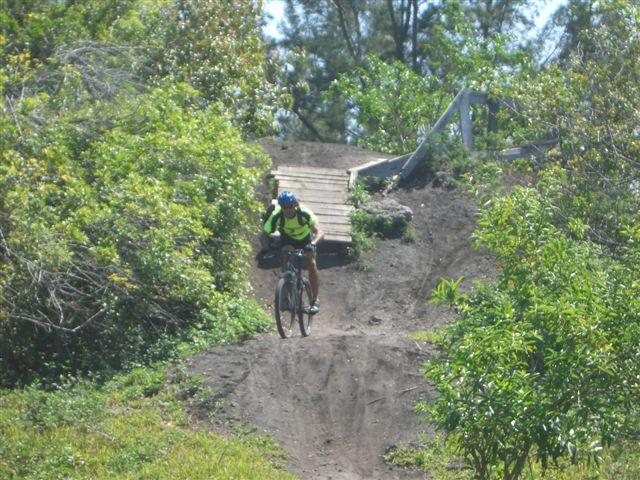 A cyclist in a bright yellow shirt and helmet rides down a dirt path with green vegetation and trees on either side. In the background, a small wooden ramp is visible, suggesting a trail designed for biking. The scene captures an outdoor biking adventure in a natural setting. Markham Park mountain bike trail.