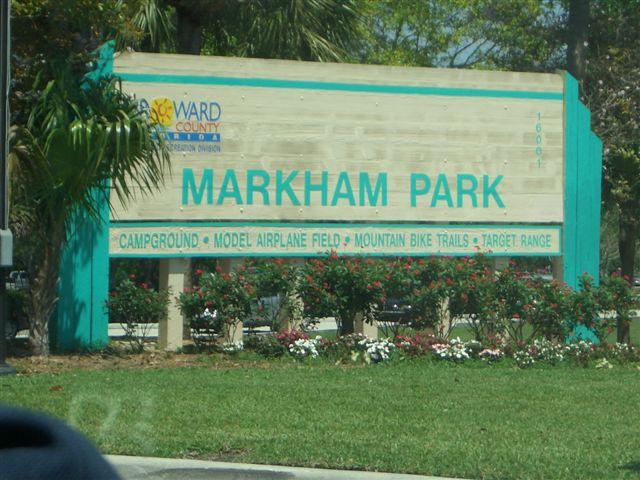Sign for Markham Park in Florida, featuring amenities such as a campground, model airplane field, mountain bike trails, and target range, surrounded by greenery and flowers. Markham Park mountain bike trail.