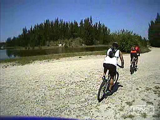 Two cyclists riding along a sandy path near a body of water, surrounded by greenery and trees under a clear blue sky. One cyclist is wearing a white shirt and black shorts, while the other is dressed in red. Markham Park mountain bike trail.