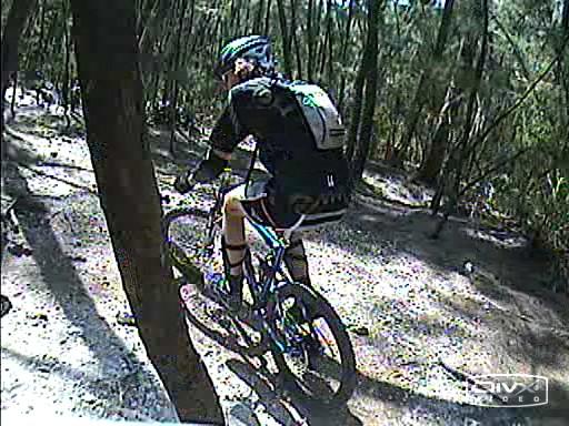 A mountain biker navigating a dirt trail in a forested area, surrounded by trees and dappled sunlight. The cyclist is wearing a helmet and sporty attire, and is focused on the path ahead. Markham Park mountain bike trail.