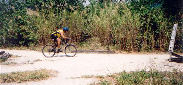 A cyclist wearing a blue helmet and a yellow jersey rides a mountain bike along a gravel path, surrounded by tall grass and greenery. A trail sign is partially visible on the right side of the image. Markham Park mountain bike trail.