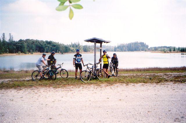Group of six cyclists taking a break by a tranquil lakeside, with trees in the background. Some are sitting on their bikes, while others stand nearby, wearing cycling gear and helmets. A small shelter is visible behind them. The scene captures a peaceful outdoor atmosphere. Markham Park mountain bike trail.
