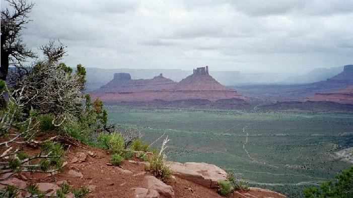 A panoramic view of a desert landscape featuring red rock formations under a cloudy sky. The foreground includes rocky terrain and scattered green vegetation, while the background showcases mesas and distant hills extending into the horizon. Porcupine Rim mountain bike trail.