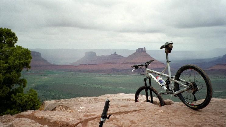 A mountain bike resting on rocky terrain, overlooking a vast landscape of red rock formations and green vegetation under a cloudy sky. Porcupine Rim mountain bike trail.