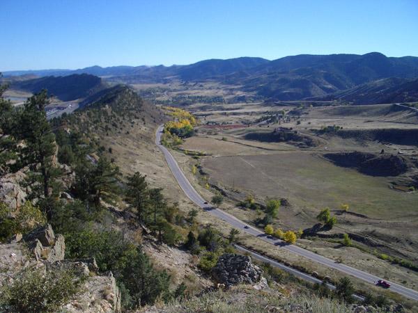 A panoramic view of a mountainous landscape featuring a winding road that cuts through open fields and valleys. The scene is marked by vegetation, including scattered trees and patches of yellow foliage, beneath a clear blue sky. The rolling hills and distant mountains create a serene and picturesque backdrop. Red Rocks / Dakota Ridge mountain bike trail.