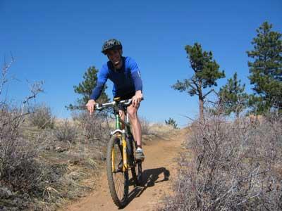 A person riding a mountain bike on a dirt trail, surrounded by sparse vegetation and trees under a clear blue sky. The cyclist is wearing a helmet and a blue shirt. Apex Park mountain bike trail.