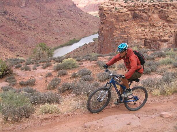 A mountain biker navigates a rugged terrain in a desert landscape, with steep rocky cliffs and a winding river visible in the background. The cyclist is wearing a blue helmet and a red jacket, riding on a bicycle along a dirt path surrounded by sparse vegetation. Porcupine Rim mountain bike trail.