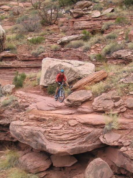 A mountain biker navigating a rocky trail surrounded by red rock formations and sparse vegetation. The cyclist is dressed in a red shirt and blue helmet, biking over uneven terrain with large boulders nearby. Porcupine Rim mountain bike trail.