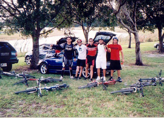 Group of five friends posing together outdoors, standing next to parked cars in a grassy area. They are wearing cycling gear and helmets, with several bicycles lying on the ground beside them. Trees and a body of water are visible in the background. Markham Park mountain bike trail.