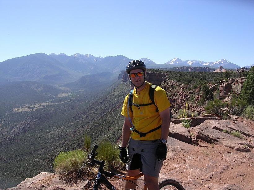 A mountain biker wearing a yellow shirt and sunglasses stands next to his bicycle on a rocky overlook, showcasing a scenic view of mountains and valleys in the background under a clear blue sky. Porcupine Rim mountain bike trail.