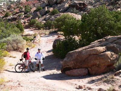 Two cyclists stand beside their mountain bikes on a rocky trail surrounded by greenery and large boulders. The landscape is rugged and natural, suggesting an outdoor adventure setting. Klondike Bluffs mountain bike trail.