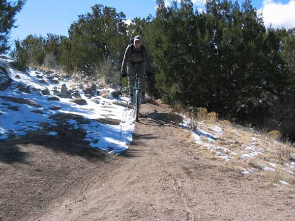 A person riding a mountain bike on a narrow dirt trail with patches of snow on the ground, surrounded by greenery and rocky terrain under a clear blue sky. Sandia Mountains Foothill Trail mountain bike trail.