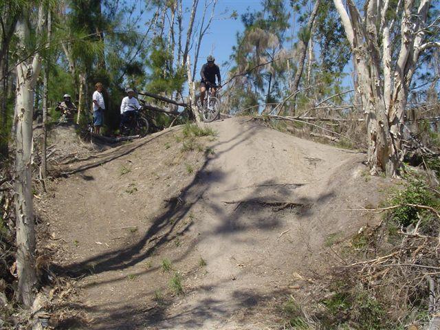 A mountain biker is captured mid-air as they jump off a dirt ramp in a forested area. In the background, a small group of spectators watches from the side, while trees and foliage create a natural setting. The terrain is sandy and slightly uneven, typical for a biking trail. Markham Park mountain bike trail.