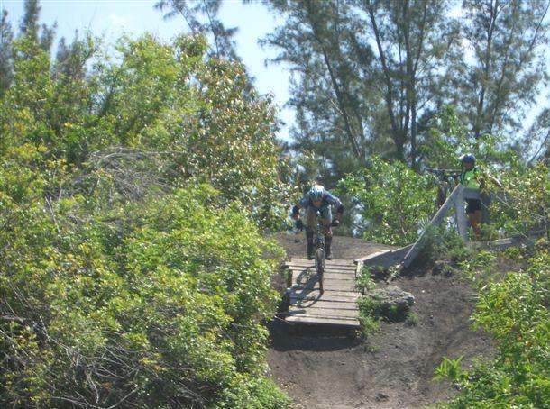 A mountain biker descends onto a wooden bridge surrounded by lush greenery, while another person stands nearby, watching the action. Trees and vibrant foliage create a natural backdrop for the biking scene. Markham Park mountain bike trail.