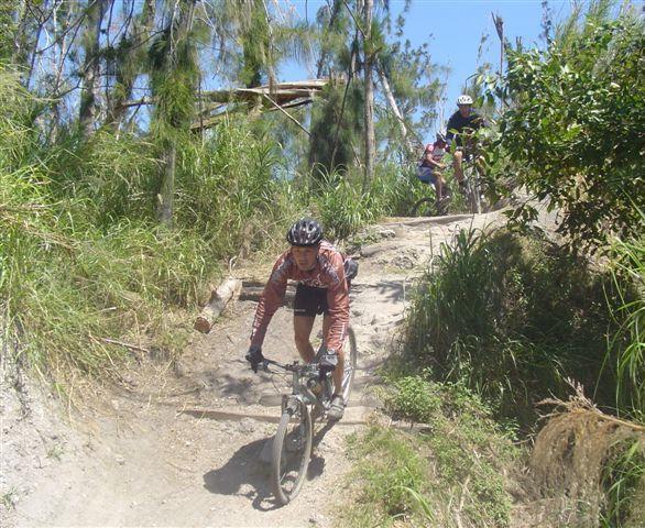 A mountain biker navigating a dirt trail surrounded by lush greenery and trees, with another cyclist visible in the background. The scene is bright and sunny, showcasing an outdoor cycling adventure. Markham Park mountain bike trail.