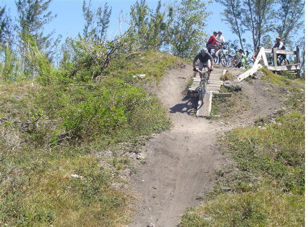 A mountain biker descends a dirt ramp on a grassy hill, surrounded by trees, while other cyclists wait at the top, enjoying the outdoors. Markham Park mountain bike trail.