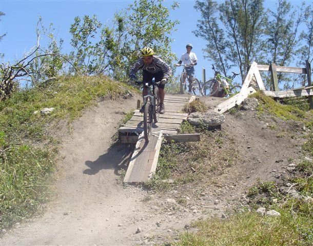 A mountain biker is descending a wooden ramp on a dirt trail, with dust kicked up behind them. In the background, another cyclist is watching, surrounded by greenery and trees under a clear blue sky. Markham Park mountain bike trail.
