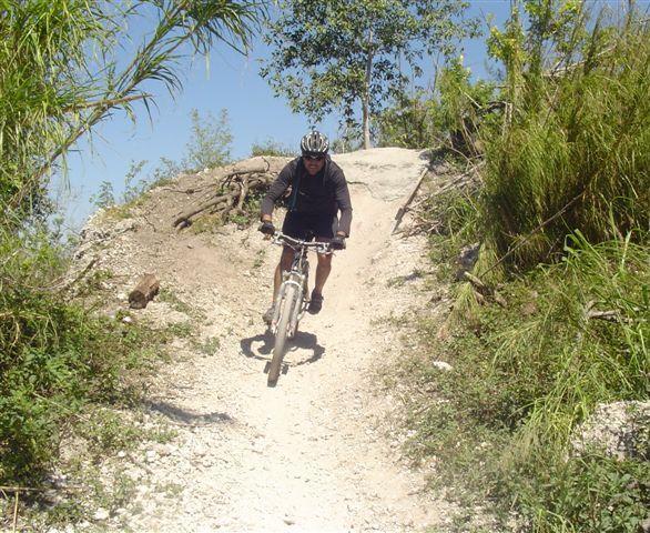 A person in a helmet rides a mountain bike down a dirt trail surrounded by greenery. The terrain is uneven and features patches of grass and small bushes. The sky is clear and blue, indicating a sunny day. Markham Park mountain bike trail.