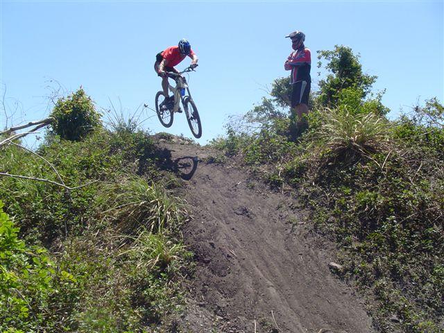 A mountain biker in a red jersey performs a jump off a dirt ramp, while another rider watches from the side. The scene is set in a natural outdoor environment with clear blue skies and greenery in the background. Markham Park mountain bike trail.
