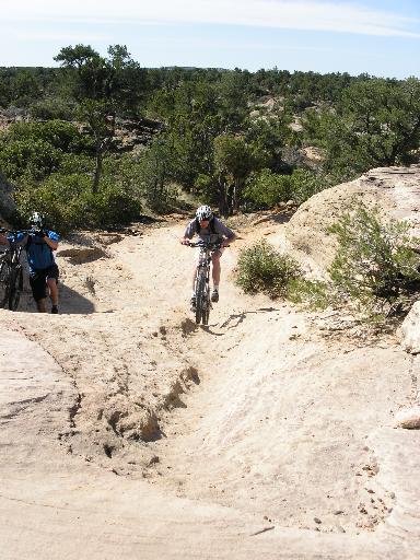 A mountain biker navigates a rocky trail, surrounded by greenery, while another cyclist stands nearby. The terrain features sandy and uneven surfaces under a clear blue sky. Gooseberry Mesa mountain bike trail.