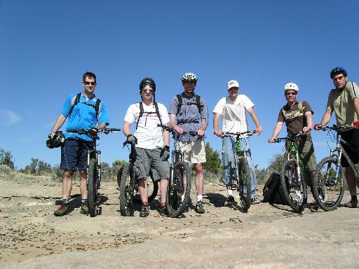 A group of six mountain bikers posed with their bicycles on a rocky trail under a clear blue sky. They are dressed in casual athletic attire and helmets, ready for an outdoor biking adventure. Gooseberry Mesa mountain bike trail.