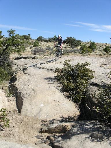 A mountain biker navigating a rocky trail under a clear blue sky. The terrain features rugged rocks and sparse vegetation, typical of a desert landscape. Gooseberry Mesa mountain bike trail.