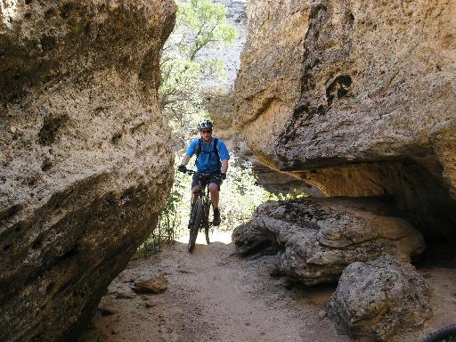 A mountain biker navigates a narrow pathway between large rock formations, with trees visible in the background. The biker wears a helmet and blue shirt, showcasing an adventurous outdoor scene. Gooseberry Mesa mountain bike trail.