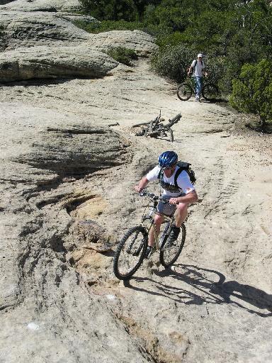A mountain biker navigating a rocky trail, wearing a blue helmet and a white shirt, with another cyclist in the background near their bike on a sunny day. Gooseberry Mesa mountain bike trail.